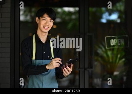 Portrait of handsome asian man waitress wearing apron and standing in front of coffee shop and smiling to camera. Stock Photo