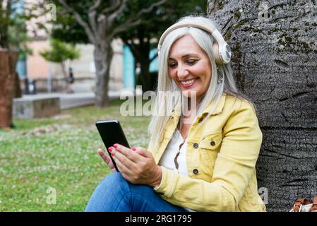 Happy modern senior female with mobile phone listening to music through headphones while sitting in green park against blurred background Stock Photo