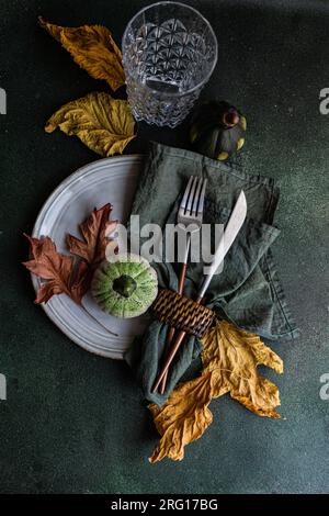 Top view of autumnal table setting with napkin, knife and fork place on plate near colorful leaves, smalls pumpkins and empty glass against dark surfa Stock Photo