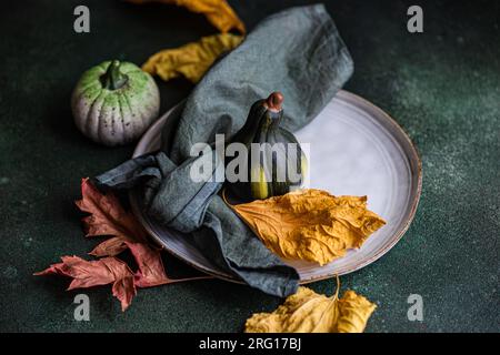 High angle of autumnal table setting with napkin, leaves and pumpkins placed on plate against dark surface Stock Photo