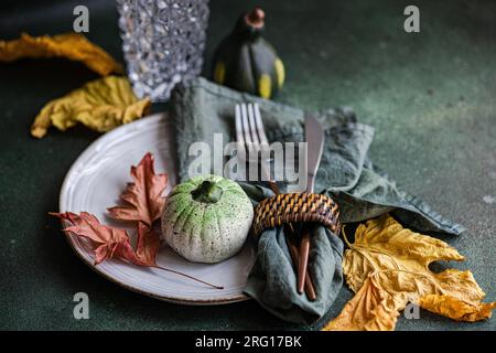 High angle of autumnal table setting with napkin, knife and fork place on plate near colorful leaves, smalls pumpkins and empty glass against dark sur Stock Photo