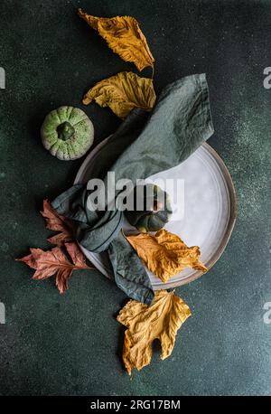Top view of autumnal table setting with napkin, leaves and pumpkins placed on plate against dark surface Stock Photo