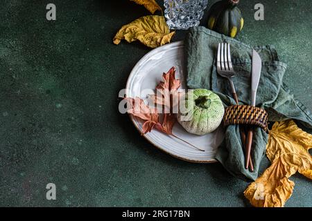 High angle of autumnal table setting with napkin, knife and fork place on plate near colorful leaves, smalls pumpkins and empty glass against dark sur Stock Photo