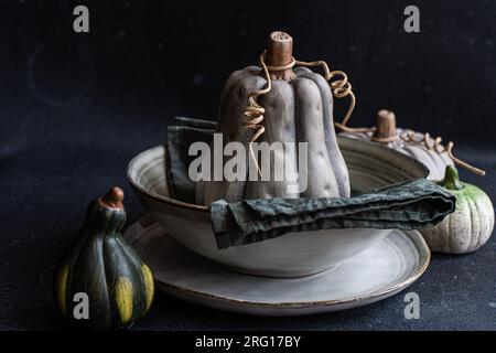 Autumnal table setting with napkin and black pumpkin placed on ceramic bowl near green pumpkins against dark surface Stock Photo