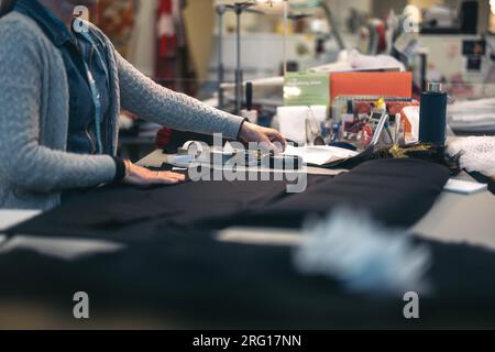 Side view of crop anonymous male fashion designer in warm jacket while standing at table with garments and working on clothes in modern design shop in Stock Photo