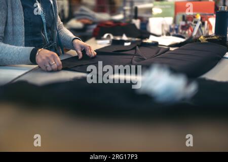 Side view of crop anonymous male fashion designer in warm jacket while standing at table with garments and working on clothes in modern design shop in Stock Photo