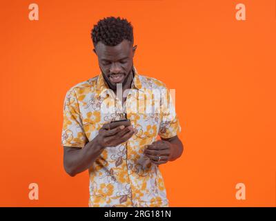 Happy young African American guy in casual shirt using smartphone while standing on orange background Stock Photo