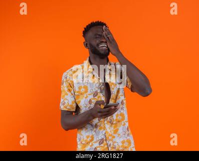 Young African American male in casual outfit with eyes closed and hand covering eye laughing while holding mobile phone against orange background Stock Photo