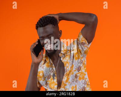 Young African American male in casual outfit looking down and laughing while having phone conversation against orange background Stock Photo