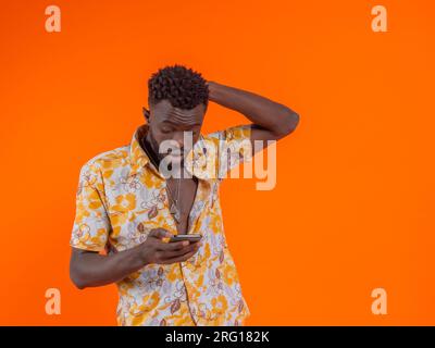 Surprised young African American male in casual outfit looking at screen of mobile phone with hand on head against orange background Stock Photo