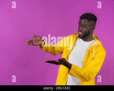 Side view of amazed young African American male looking away while standing with open palms and pointing with hands against purple background Stock Photo