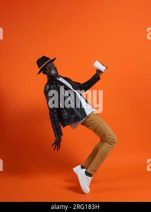 Full body side view of confident happy African American male in stylish leather outfit and hat with soda in hip hop dance against orange background Stock Photo