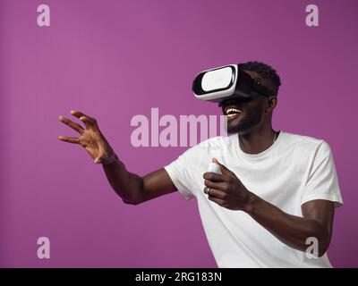 Excited young African American male in casual clothes and virtual reality headset while standing with half folded fingers of raised hand using control Stock Photo