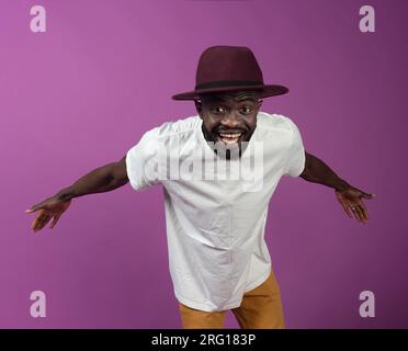 Excited young bearded African American male in white shirt and hat smiling and looking at camera while standing with outstretched hands behind and ben Stock Photo