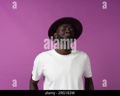 Exhausted young African American male in hat and white shirt with eyes closed while standing alone in depressed and grief mood against purple backdrop Stock Photo