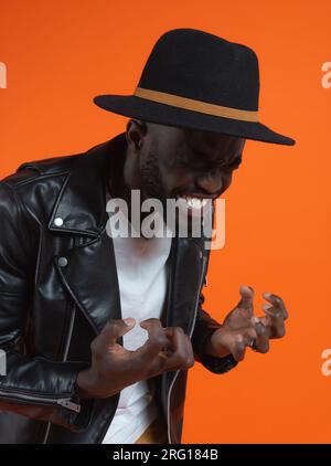 Portrait of angry African American bearded male in stylish leather jacket and hat making angry face with claws while standing against orange backgroun Stock Photo