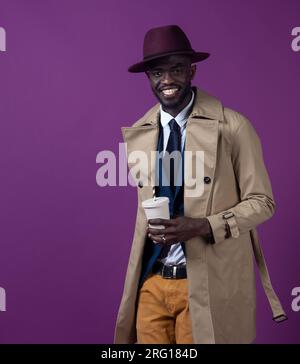Smiling young African American male in stylish coat tie and hat looking at camera while standing with takeaway cup near purple backdrop Stock Photo