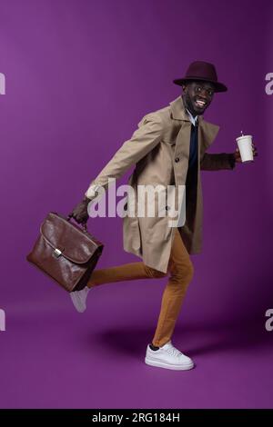 Full body side view of smiling young African American male in hat looking at camera while running with leather briefcase and takeaway cup with straw a Stock Photo