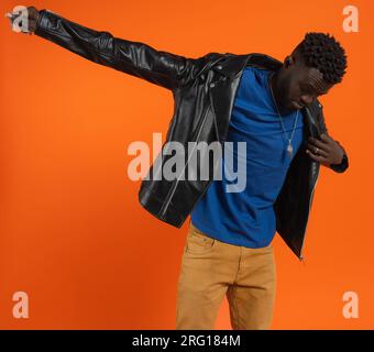 Young African American male with dark curly hair looking down while standing stretching arm and wearing black leather jacket against orange background Stock Photo