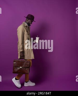 Side view of smiling young African American male in hat looking down while walking with leather briefcase and takeaway cup with straw against purple b Stock Photo