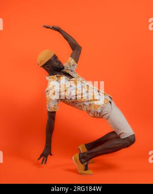 Full body side view of young African American male in orange printed short sleeves shirt and cap looking away while raising arm bending back on toes a Stock Photo