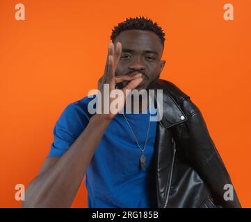 Portrait of confident young African American male with necklace, short curly hair looking at camera while standing with leather jacket and showing OK Stock Photo