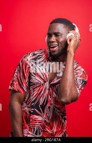 Positive African American male in casual clothe shirt listening to music and enjoying songs with wireless earphones against red background Stock Photo