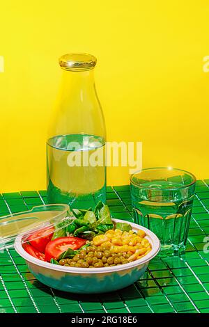 Salad bowl with slices of tomato, spinach leaves, corn kernels and peas placed on green surface near bottle and glass of water against yellow wall Stock Photo