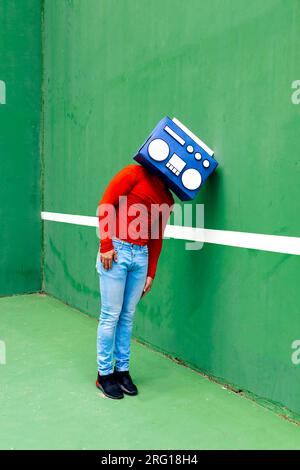 Upset anonymous male in casual clothes covering face with blue boombox leading on green wall Stock Photo