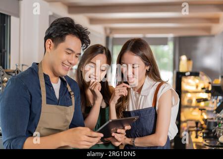 Three barista entrepreneur team meeting at coffee shop. Young smiling Asian male and female cafe employees having discussion on work and menu with tablet computer Stock Photo