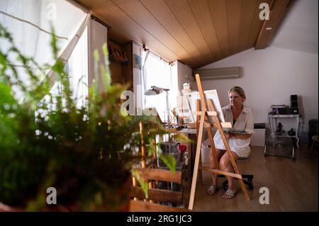 Interior of a studio with a senior woman painting an oil painting. Stock Photo
