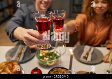 Focus on two glasses of red wine held by young cheering man and woman sitting by table served with homemade food prepared for guests Stock Photo