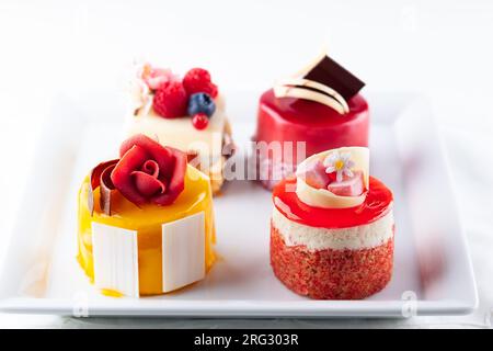 Various mini cakes on a white plate. Sweets decorated with fresh berries and flowers for holiday. Stock Photo