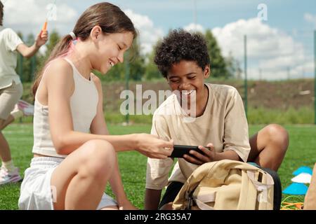 Two laughing kids in activewear watching funny and curious online video on screen of smartphone held by AFrican AMerican schoolboy Stock Photo