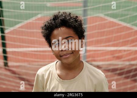 Happy cute African American schoolboy looking at camera against goal net while standing on sports ground or stadium on sunny day Stock Photo