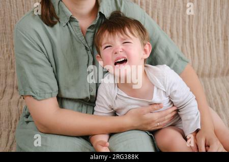 Portrait of an adult woman with a crying baby sitting on a sofa, female age 37 years and male two years Stock Photo