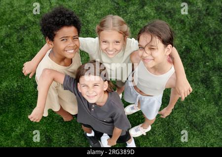 Above angle of several happy intercultural children in activewear standing against green lawn or football field and looking at camera with smiles Stock Photo