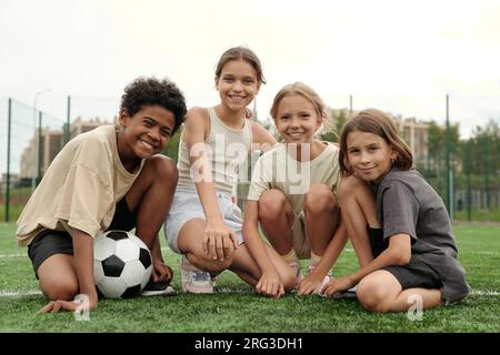 Team of four active friends and football players looking at camera with smiles while sitting on green lawn of football field at stadium Stock Photo