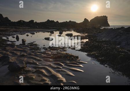 Sunset over a shore at low tide, showing rippled sand, a rock pool and rocks, at Hartland Quay, Hartland, Atlantic coast of Devon, Great Britain. Stock Photo