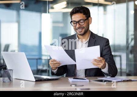 Successful mature experienced financier on paper work, Arab smiling satisfied with results of financial achievement, reviewing contracts, invoices and statements with accounts, businessman in suit. Stock Photo