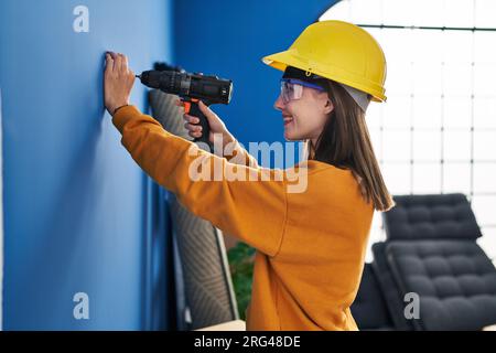 Young blonde woman wearing hardhat using dirll on wall at new home Stock Photo