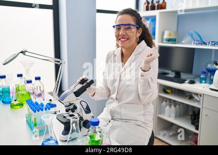 Young hispanic woman working at scientist laboratory screaming proud, celebrating victory and success very excited with raised arm Stock Photo