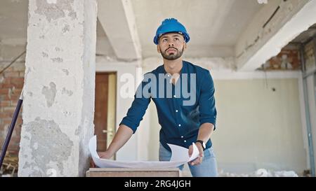 Young hispanic man architect wearing hardhat looking at blueprints at construction site Stock Photo