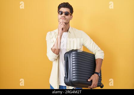 Young hispanic man holding suitcase going on summer vacation thinking concentrated about doubt with finger on chin and looking up wondering Stock Photo