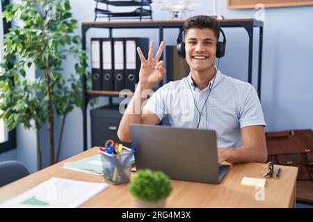 Young hispanic man working at the office wearing headphones showing and pointing up with fingers number three while smiling confident and happy. Stock Photo