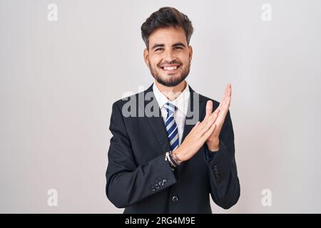 Young hispanic man with tattoos wearing business suit and tie clapping and applauding happy and joyful, smiling proud hands together Stock Photo