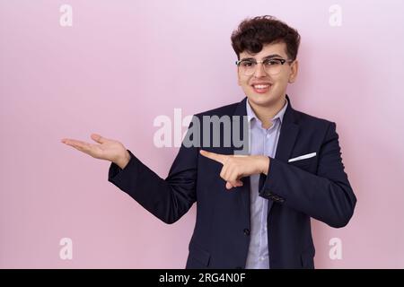 Young non binary man with beard wearing suit and tie amazed and smiling to the camera while presenting with hand and pointing with finger. Stock Photo
