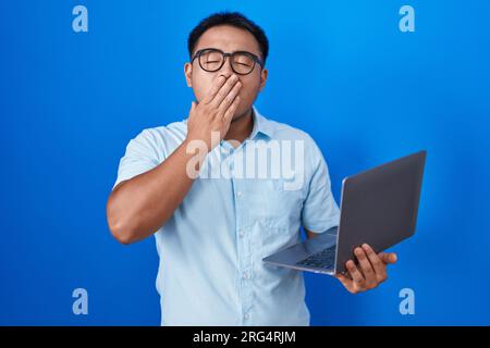Chinese young man using computer laptop bored yawning tired covering mouth with hand. restless and sleepiness. Stock Photo