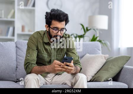 Serious thinking man sitting on sofa in living room at home, Indian man reading news on phone, using app on smartphone, browsing internet pages. Stock Photo