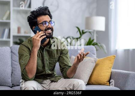 Young successful and smiling man talking on the phone while sitting on the sofa in the living room, Indian cheerfully chatting with colleagues and friends. Stock Photo